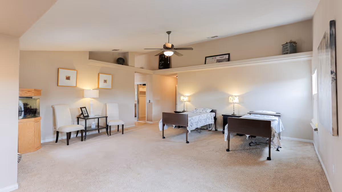A spacious senior care bedroom with two hospital-style beds, each with a bedside table and lamp. The room has beige walls and carpet, a ceiling fan with light, two white chairs with a small table between them, and a fish tank in the corner. There are framed pictures on the walls and a large window letting in natural light.