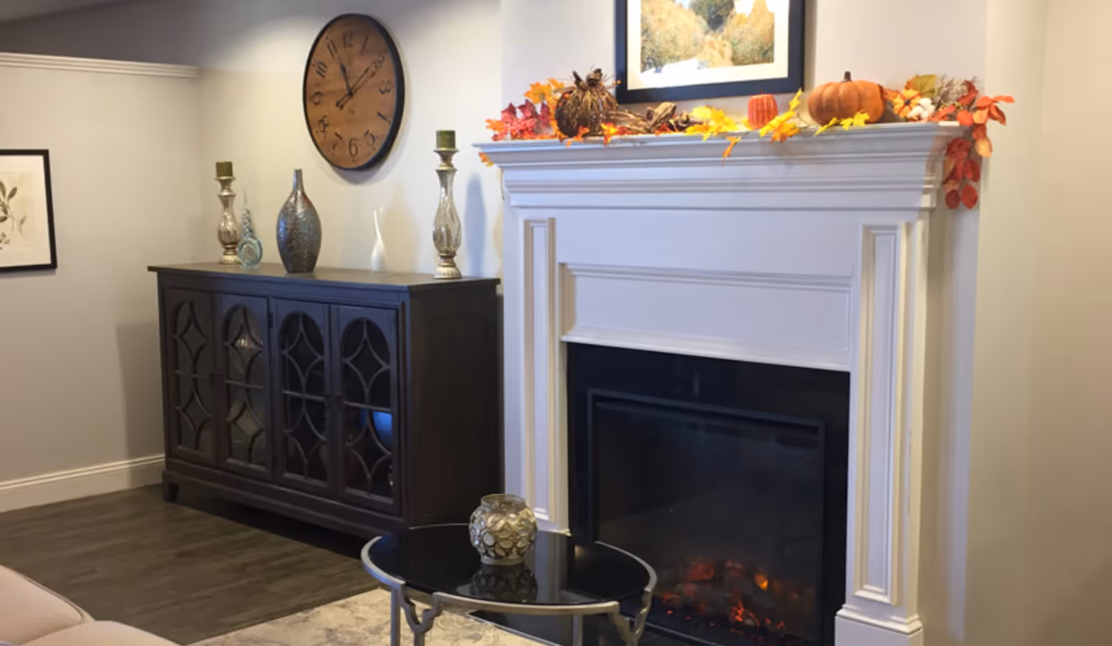 Interior living room showing a white decorative fireplace with fall mantel decor, a dark wood cabinet with vases, a round wall clock, and a glass-top coffee table.