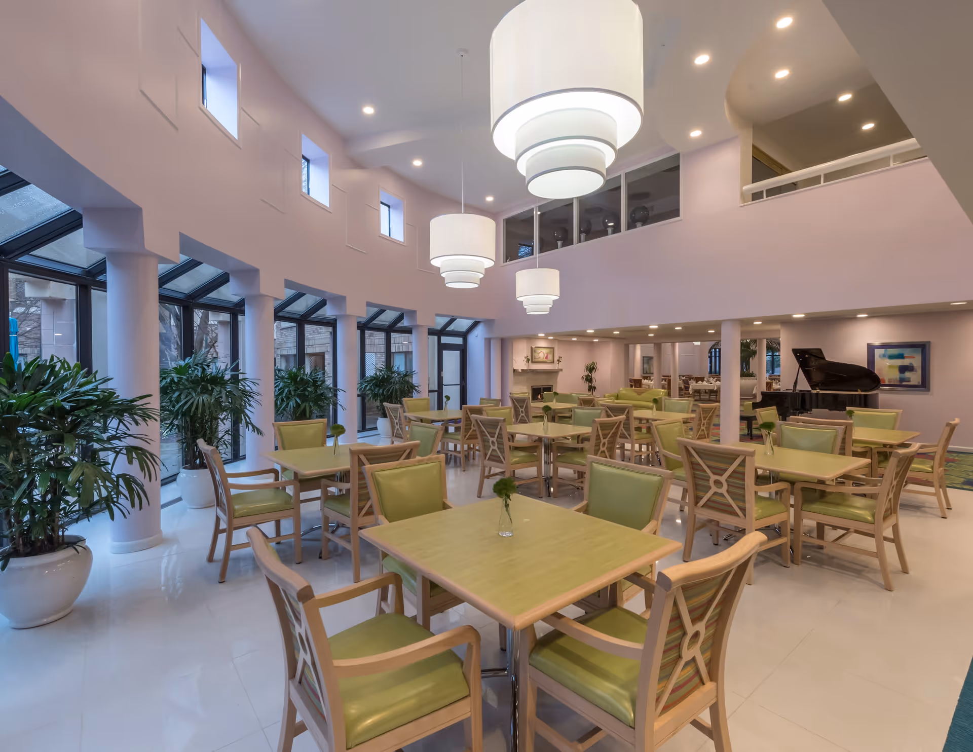A spacious senior living dining room with multiple light green tables and matching chairs arranged neatly. Large windows with black frames line one side, allowing natural light to enter. Several large white pendant lights hang from the high ceiling. There are potted plants along the windows and a black grand piano in the background near a fireplace and artwork on the walls.