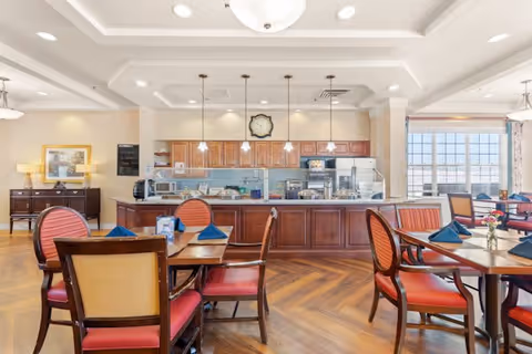 A bright dining area with wooden tables and red cushioned chairs, each table set with blue napkins and small flower vases. In the background, there is a kitchen counter with wooden cabinets, a clock on the wall, and pendant lights hanging from the ceiling. Large windows on the right side let in natural light.