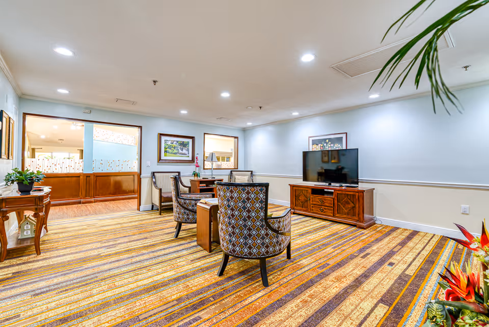 Bright common living area with patterned chairs around a small table, a TV on a wooden console, and a colorful striped carpet.