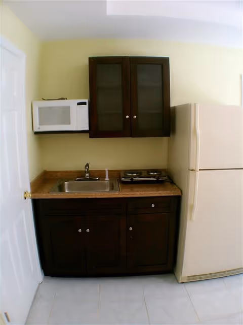 Small kitchen area with a white microwave mounted on the wall, dark wooden cabinets above and below a countertop with a sink and a two-burner electric stove. A beige refrigerator is positioned to the right of the countertop. The walls are light yellow and the floor is tiled in white.