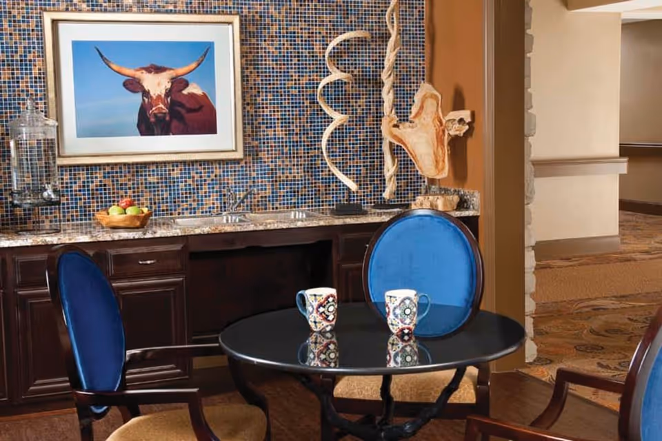 A small seating area with a round black table and three chairs with blue cushions. Two colorful mugs are placed on the table. Behind the table is a countertop with a sink, a bowl of fruit, and decorative wooden sculptures. The wall behind the countertop is covered with small mosaic tiles and features a framed picture of a longhorn cow.