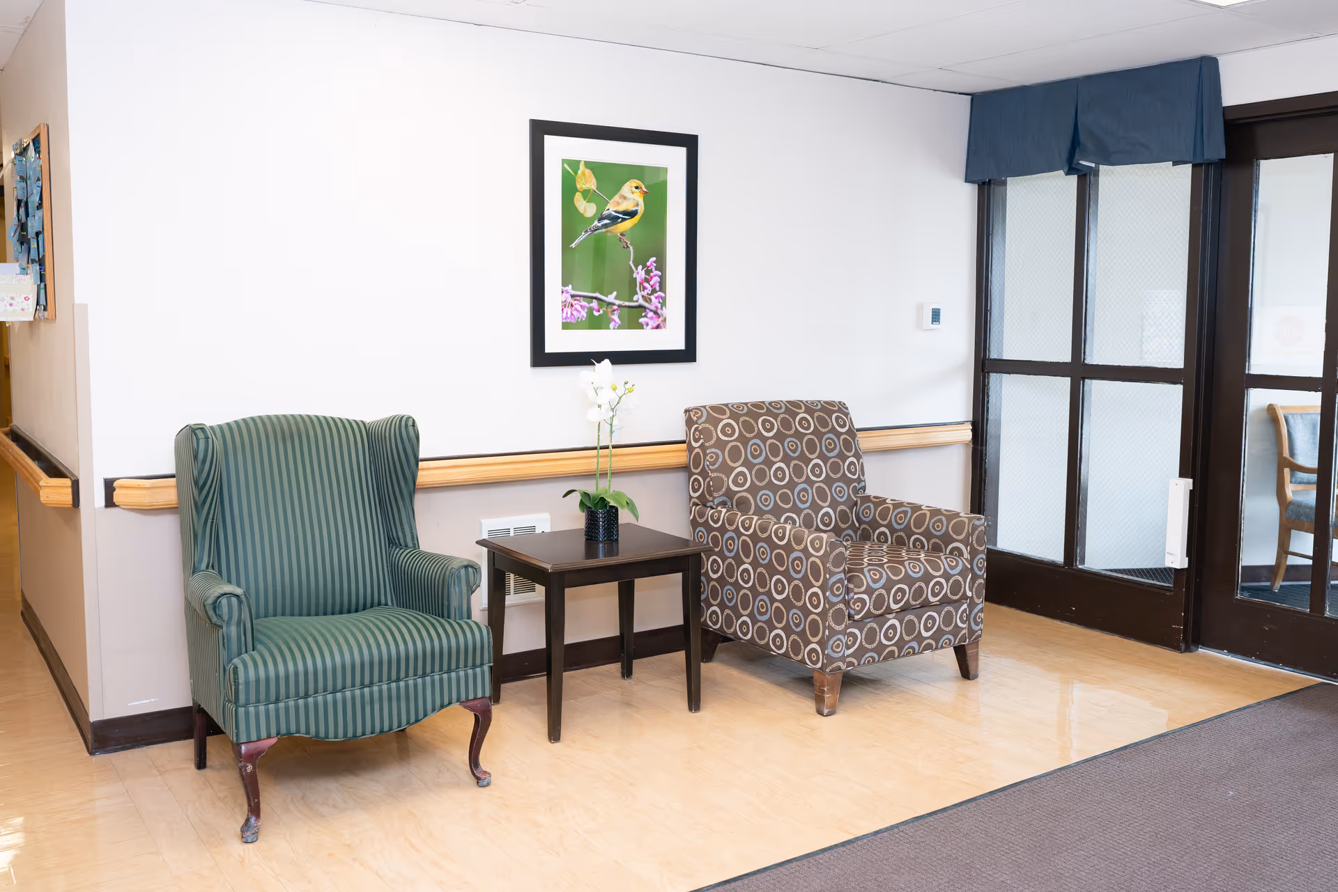 Two upholstered armchairs flanking a small side table with a plant in a bright seating area by glass doors and a framed bird picture on the wall.