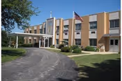 Exterior view of Misericordia Nursing & Rehabilitation Center, a two-story brick building with multiple windows, an American flag on a flagpole near the entrance, and a curved driveway leading to the main entrance under a covered drop-off area.