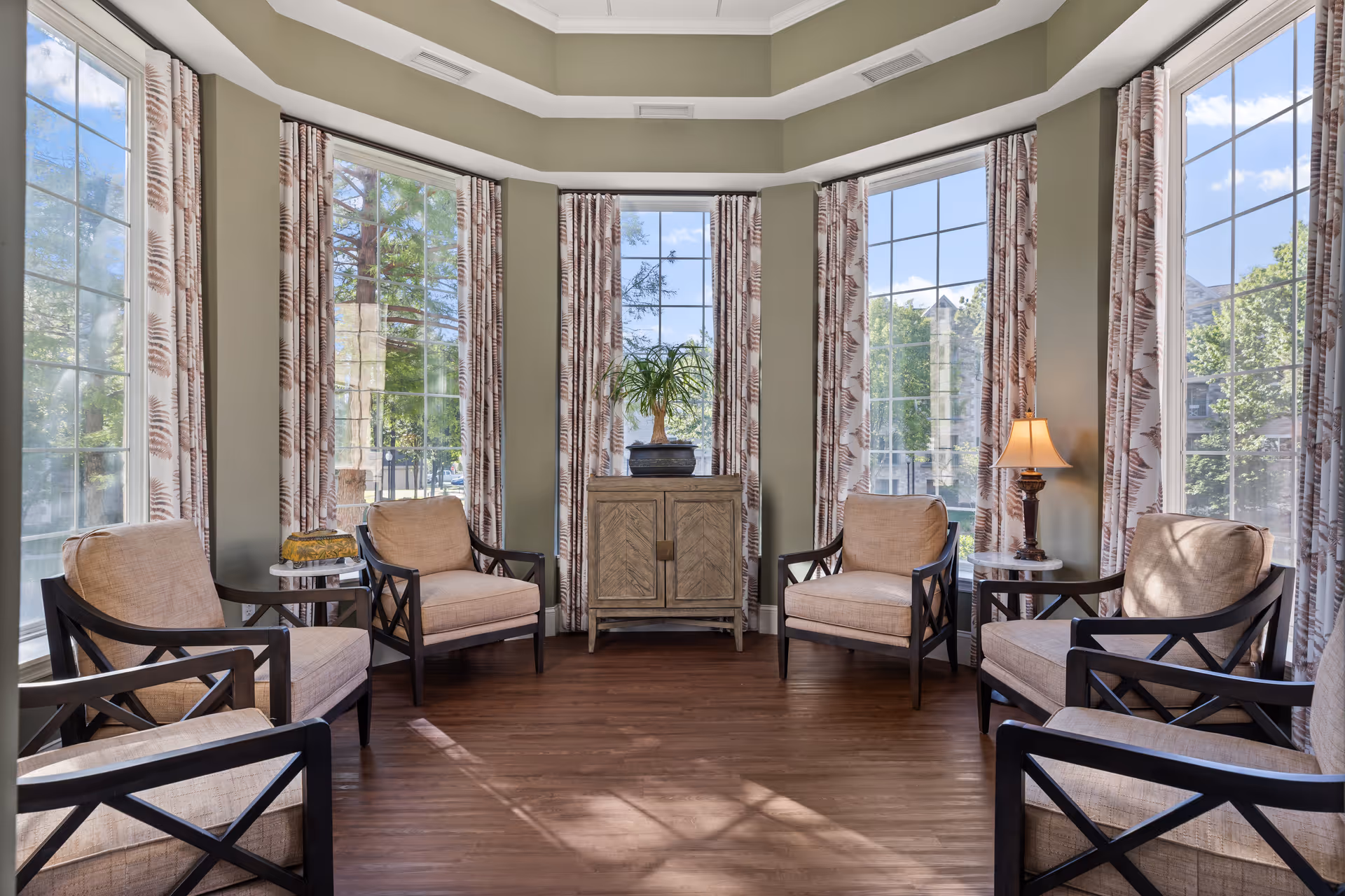 Sunlit lounge with beige armchairs arranged in a semicircle in front of tall bay windows and a small cabinet topped with a plant.