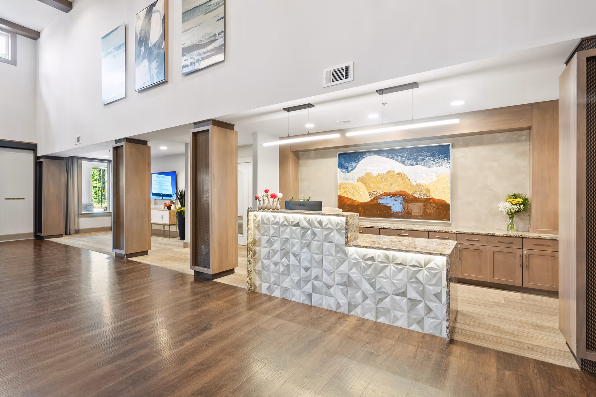 Bright modern reception area with a textured front desk, wooden pillars, artwork, and cabinetry in a senior living facility.