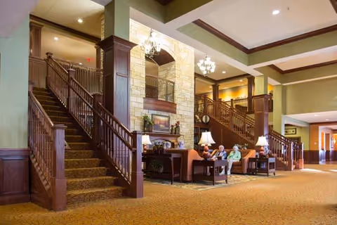 Spacious senior living facility common area with carpeted floors, wooden staircases on both sides, a stone accent wall with a painting, chandeliers, and several seniors seated on sofas and chairs around a coffee table.