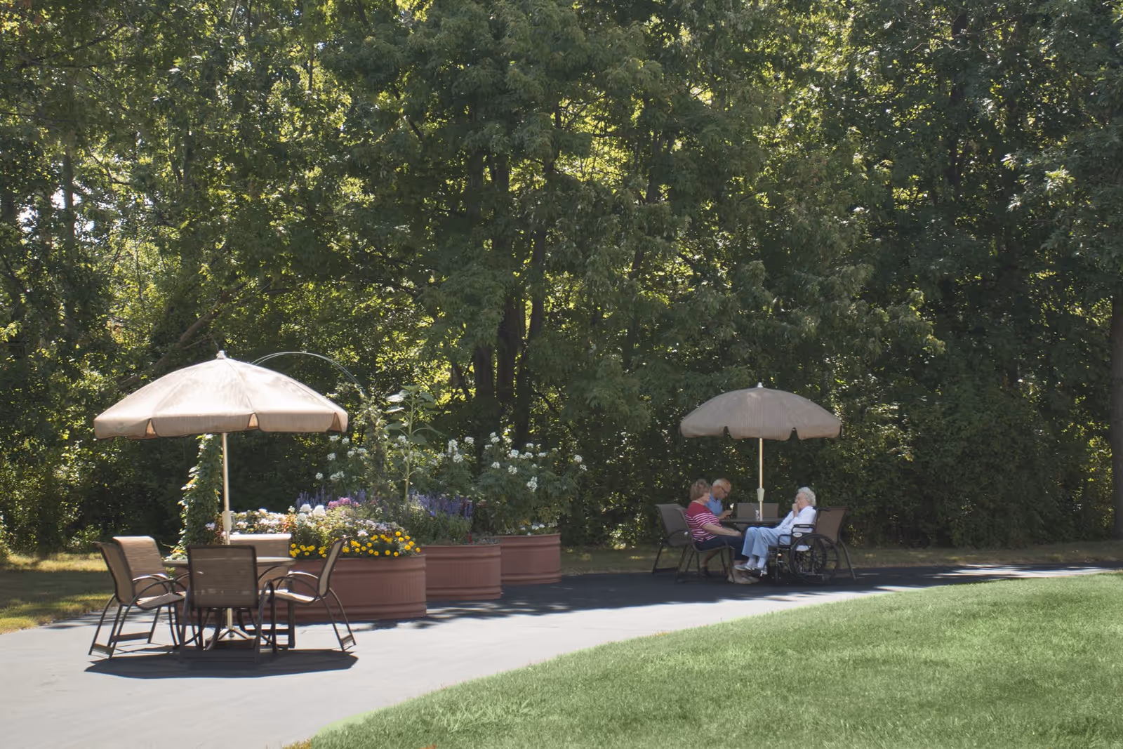Outdoor seating area with two tables, each shaded by a large beige umbrella. One table is empty with four chairs around it, while the other table has three elderly people sitting and conversing, one of whom is in a wheelchair. The area is surrounded by lush green trees and large flower planters with colorful blooms.