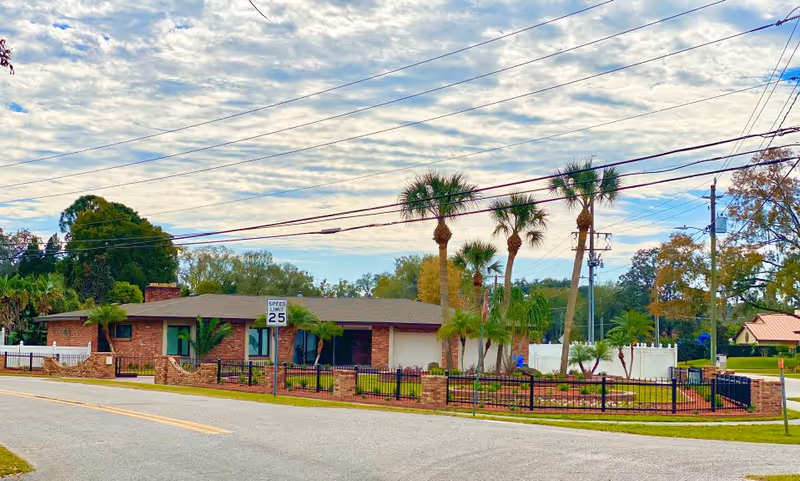 Exterior view of a single-story brick building with a low-pitched roof, surrounded by a black metal fence with brick pillars. The property features several palm trees and a well-maintained lawn. A speed limit sign indicating 25 mph is visible near the street in front of the building. The sky is partly cloudy with power lines crossing overhead.