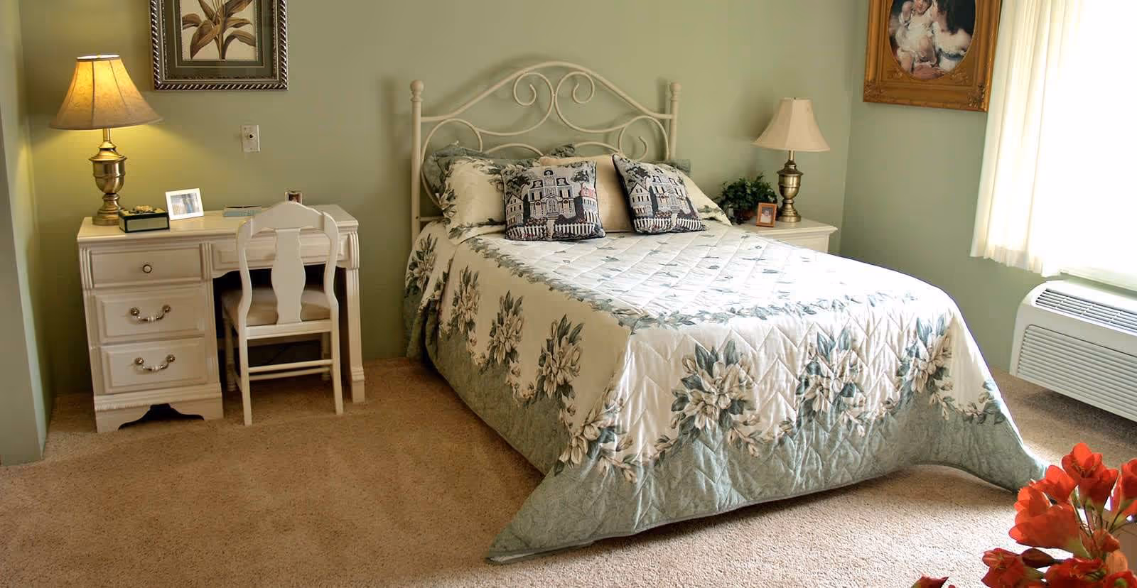 A cozy bedroom with a double bed covered in a floral quilt and two decorative pillows. There is a white desk with a chair and a lamp on the left side of the bed, and a nightstand with a lamp and a small plant on the right side. A framed picture hangs on the wall above the desk, and a window with sheer curtains lets in natural light on the right side of the room. The carpet is beige, and there is a heating/cooling unit below the window.