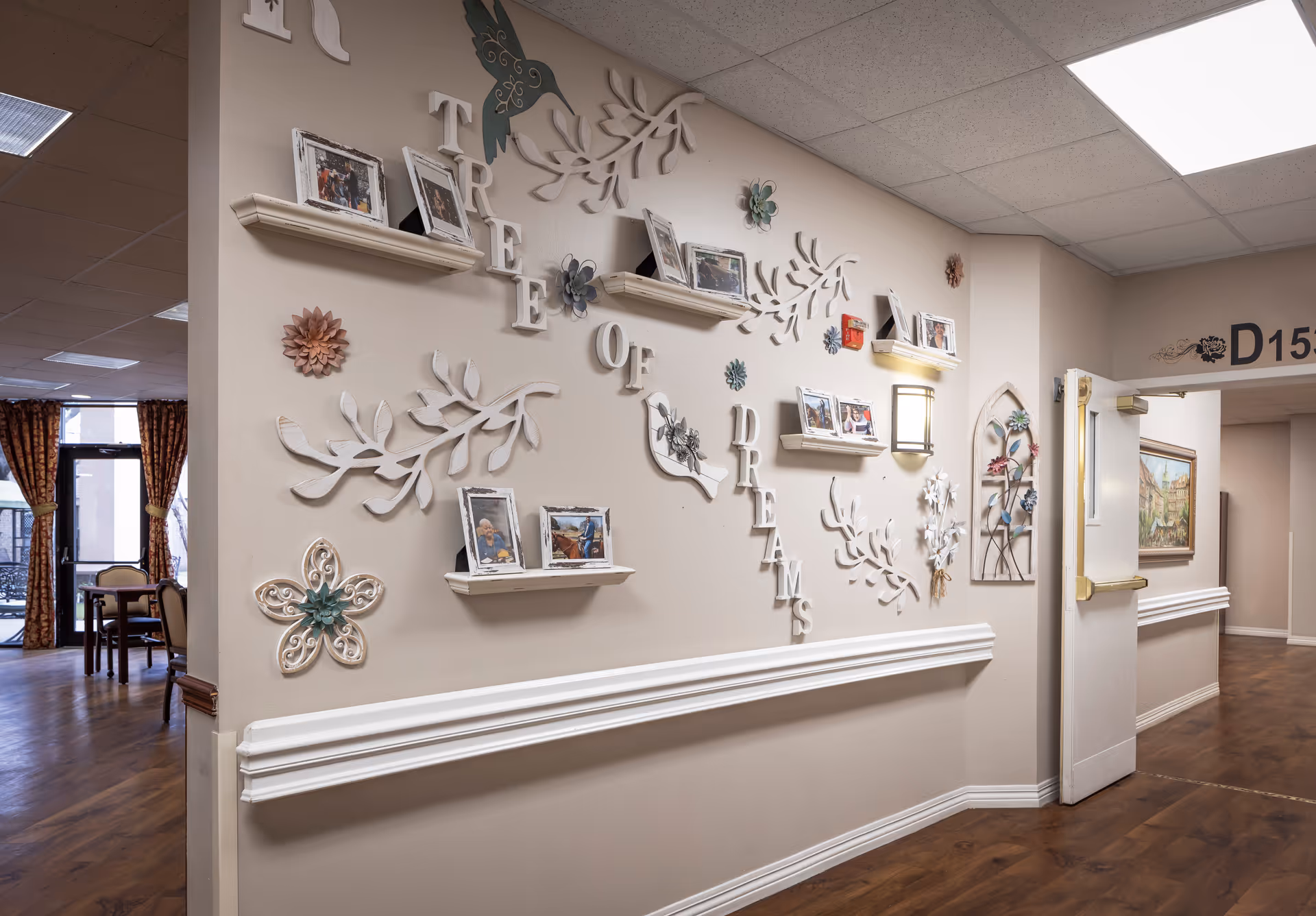 Interior hallway of an assisted living facility with decorative wall art featuring the words 'Tree of Dreams' and several small photo frames on shelves. The hallway has wooden flooring, beige walls, and a door labeled D15. In the background, a dining area with tables, chairs, and large windows with curtains is visible.