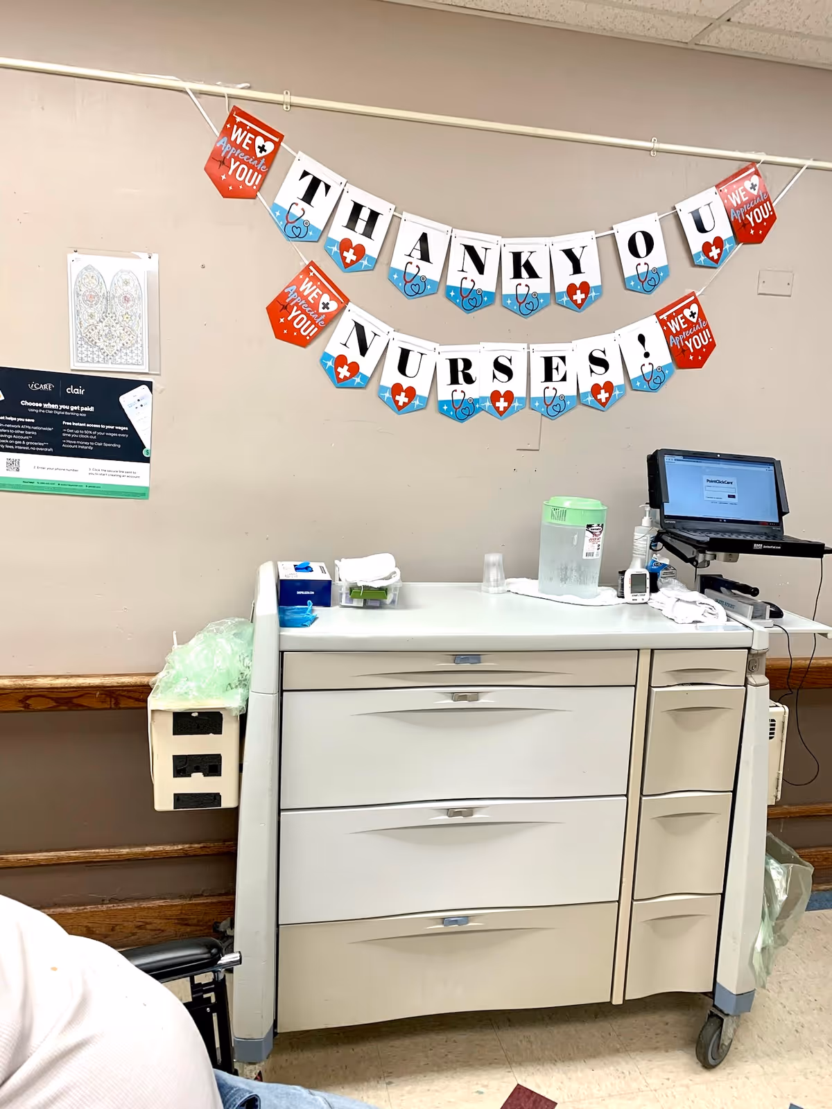 A medical supply cart and laptop beneath a wall banner reading 'Thank You Nurses!' in a facility room.