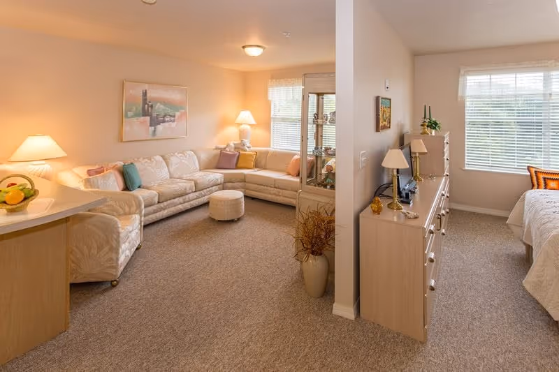 A cozy living area with a large beige sectional sofa adorned with colorful throw pillows, a small round ottoman, and two table lamps providing warm lighting. Adjacent to the living area is a bedroom with a bed, dresser, and large windows letting in natural light. The space is carpeted and decorated with framed artwork and plants.