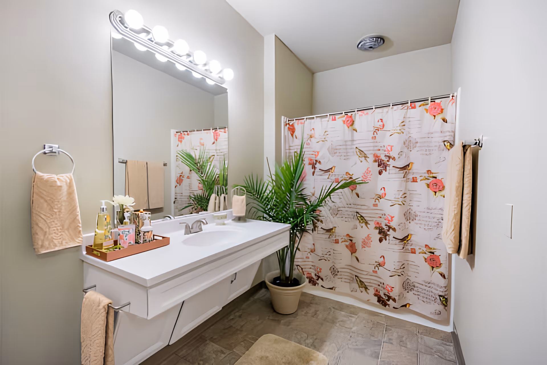A clean and bright bathroom featuring a white vanity with a large mirror above it, illuminated by a row of round lights. On the vanity countertop, there is a tray with various toiletries and a small vase with white flowers. A beige towel hangs on a ring to the left and another beige towel hangs on a bar to the right. A potted green plant is placed on the floor next to the shower area, which has a decorative shower curtain with floral and bird patterns. The floor is tiled in a neutral tone.