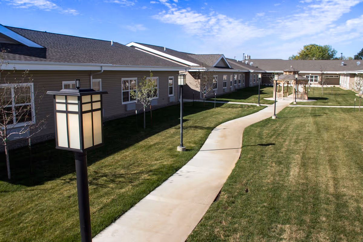 Curving concrete walkway through a grassy courtyard with lamp posts, a gazebo, and a single-story assisted living building.