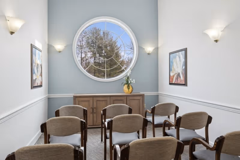 A small meeting or chapel room with two rows of beige cushioned chairs facing a wooden cabinet under a large round window. The walls are painted white and light blue with two floral paintings and wall-mounted lights.