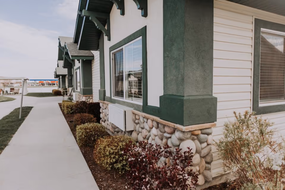 Exterior view of a building with green and white walls, stone accents at the base, windows with blinds, and a sidewalk alongside landscaped bushes and plants.