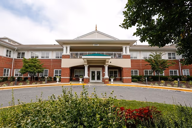 Front exterior view of a two-story senior living facility building with red brick and white siding, a covered entrance with columns, and landscaped greenery in the foreground.