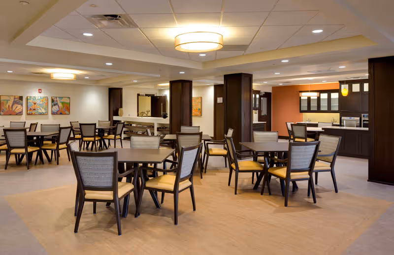 Spacious communal dining area with multiple square tables and upholstered chairs, warm lighting, and a kitchenette/bar in the background.
