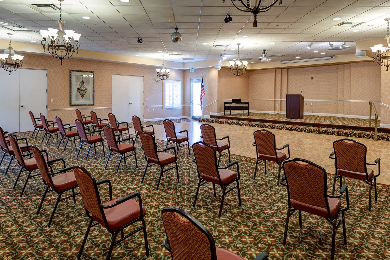 A large meeting or event room with rows of red cushioned chairs spaced apart on a patterned carpet. The room features a small stage with a podium and a keyboard, chandeliers hanging from the ceiling, and an American flag near a door with windows.