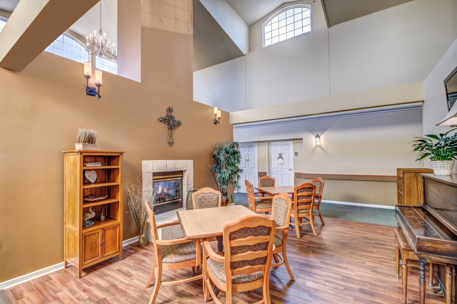 A bright communal lounge with wooden tables and chairs, a fireplace, piano, bookshelf, and tall arched windows under a high ceiling.