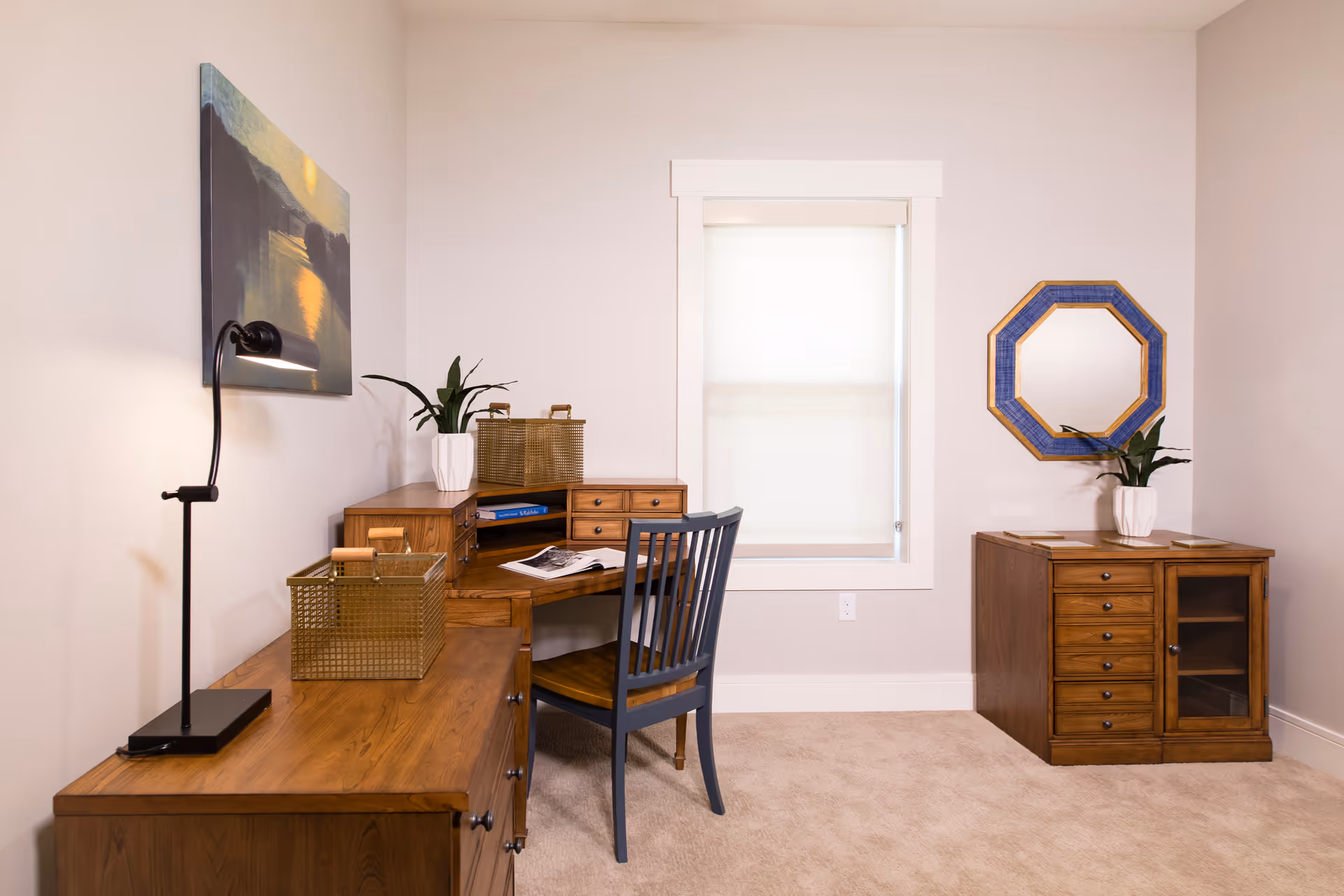 A small office or study area with wooden furniture including a desk, a chair, and a cabinet. The desk has a black desk lamp, two decorative baskets, a plant in a white pot, and an open book. There is a window with a white blind in the center of the wall and a blue octagonal mirror hanging on the right wall above the cabinet. The walls are light-colored and the floor is carpeted.