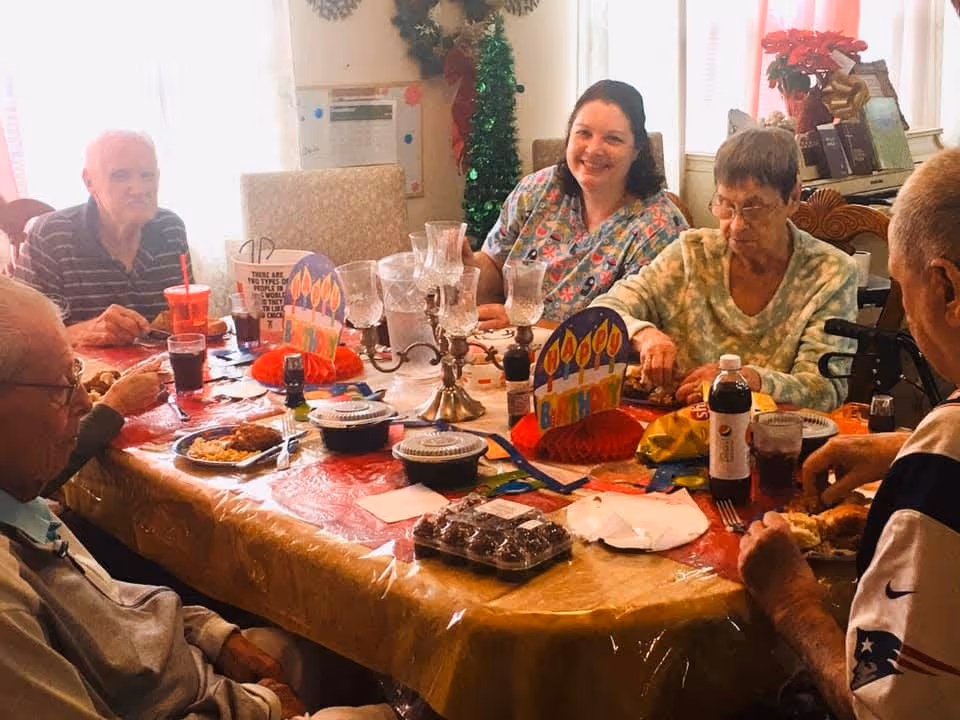 A group of elderly people and a caregiver sitting around a dining table enjoying a meal together. The table is decorated with birthday-themed centerpieces and covered with a plastic tablecloth. There are plates of food, drinks, and desserts on the table. The room has a festive atmosphere with a wreath and decorations in the background.
