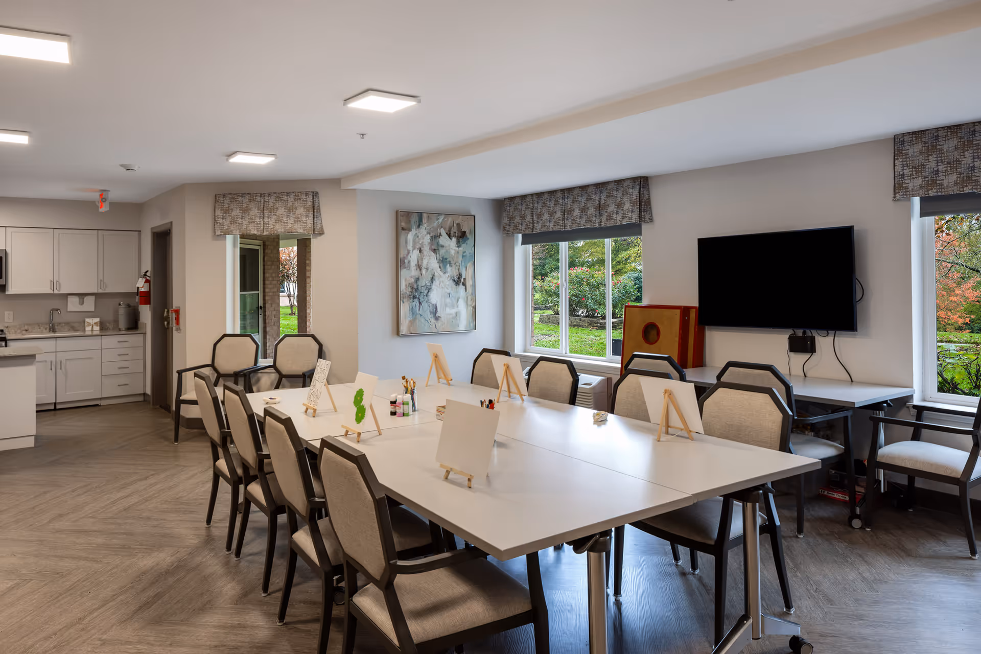 A bright and spacious activity room with a large white table surrounded by cushioned chairs. The table has small easels with blank canvases and art supplies like paint bottles and brushes. The room has large windows with patterned valances, letting in natural light and showing greenery outside. A flat-screen TV is mounted on the wall, and there is a kitchenette area with cabinets and a countertop in the background.