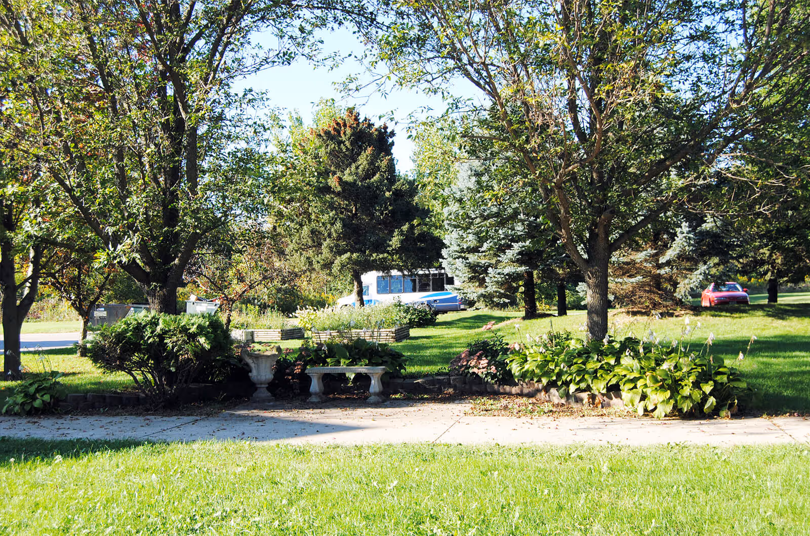 Grassy front yard with trees, a stone bench and planters, and parked cars visible in the background.