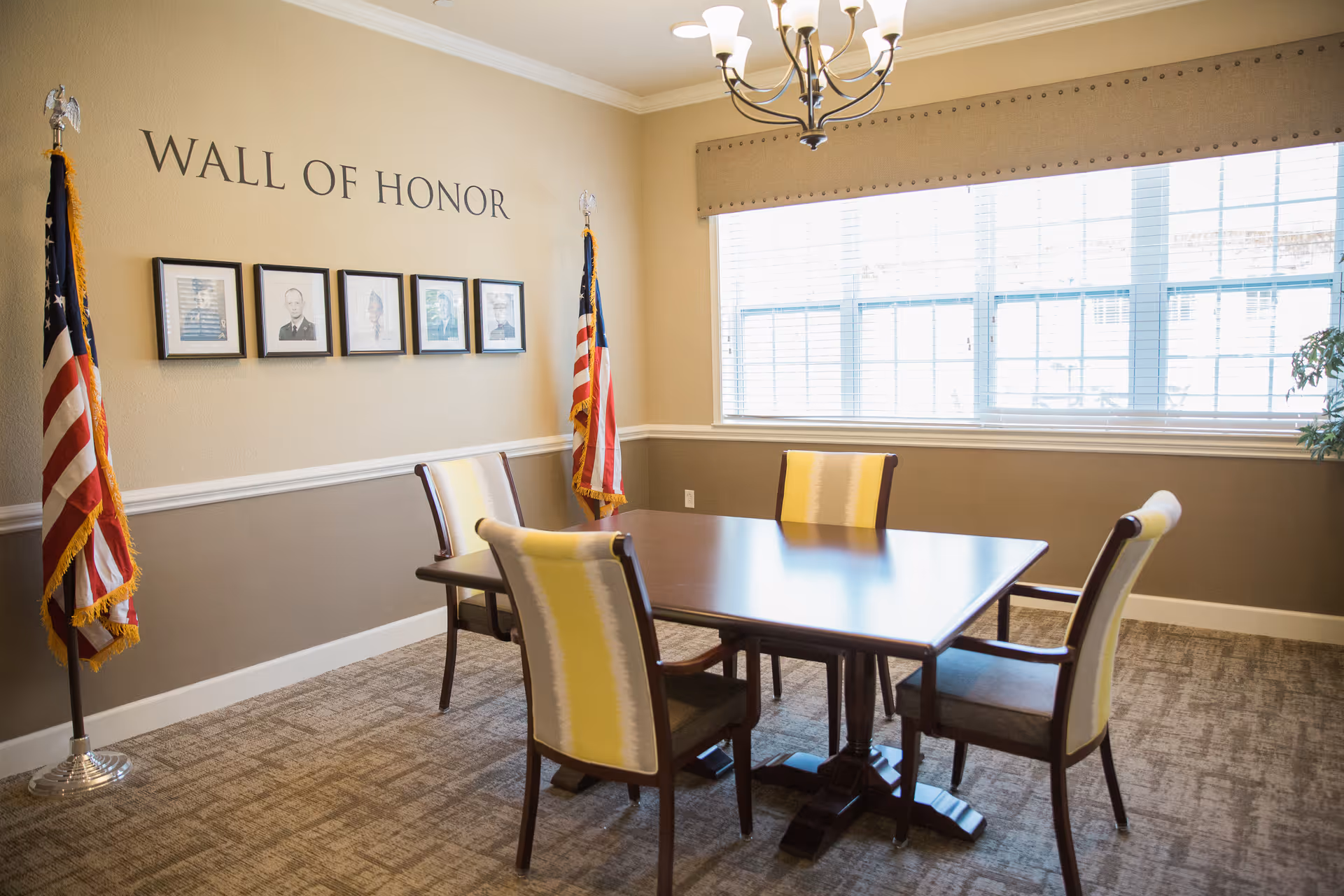 A small meeting or dining room with a rectangular wooden table and four chairs with yellow and white upholstery. On the wall behind the table is a 'Wall of Honor' display featuring five framed photographs and two American flags on stands. A large window with blinds allows natural light into the room, and a chandelier hangs from the ceiling.