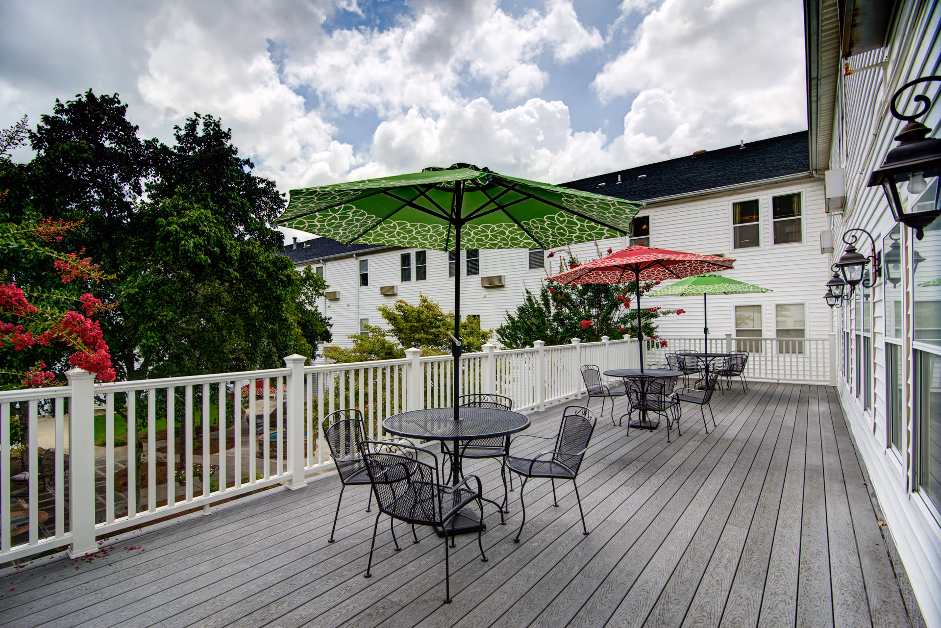 Outdoor deck area at Morningside of Vestavia Hills with several black metal tables and chairs. Each table has a colorful umbrella, including green and red ones. The deck is surrounded by a white railing and overlooks trees and other buildings under a partly cloudy sky.