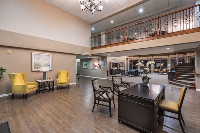 Spacious senior living facility common area with wooden flooring, two yellow armchairs with a small table and lamp between them on the left, a black desk with a yellow chair and two patterned chairs in the center, and a staircase leading to an upper level with decorative railing. The area is well-lit with ceiling lights and a chandelier.