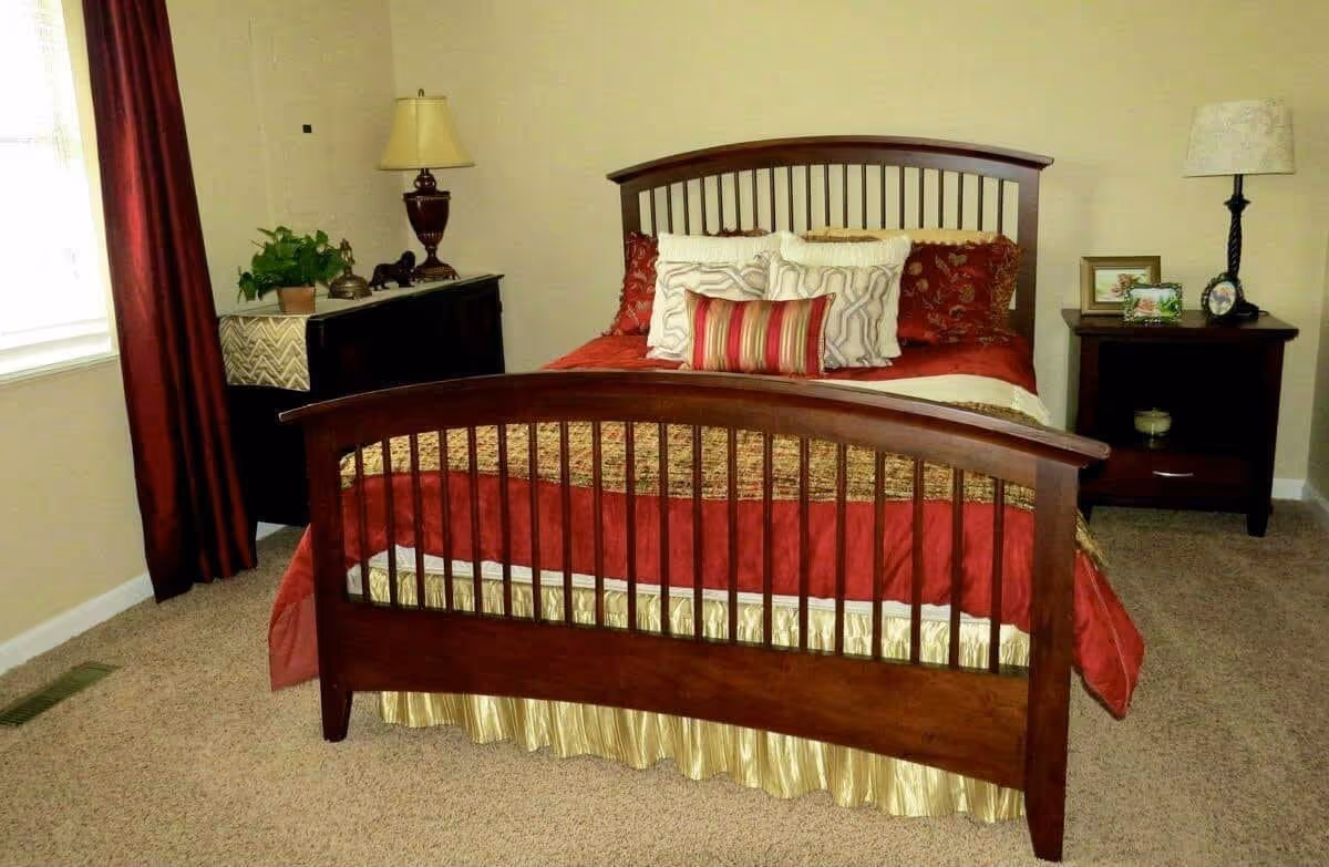 A bedroom with a wooden bed frame featuring vertical slats on the headboard and footboard. The bed is made with red and gold bedding and multiple decorative pillows. On either side of the bed are dark wooden nightstands with lamps, framed photos, and decorative items. A window with red curtains is visible on the left side of the room, and the floor is carpeted.