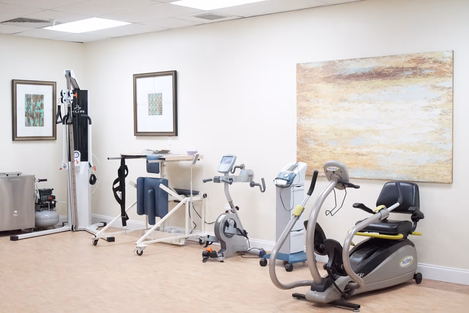 A small exercise room with various fitness equipment including a recumbent exercise bike, a stationary bike, and other physical therapy machines. The room has light-colored walls adorned with framed artwork and a large abstract painting. The floor is wooden, and the ceiling has white tiles.