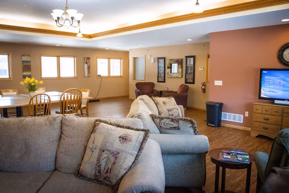 A cozy living room area with a beige sectional sofa adorned with patterned cushions. In the background, there is a dining area with wooden chairs and a table with a vase of yellow flowers. The room has wooden flooring, beige walls, and several windows letting in natural light. A TV is mounted on a wooden dresser on the right side, and two purple armchairs are placed near the back wall with framed artwork above them.