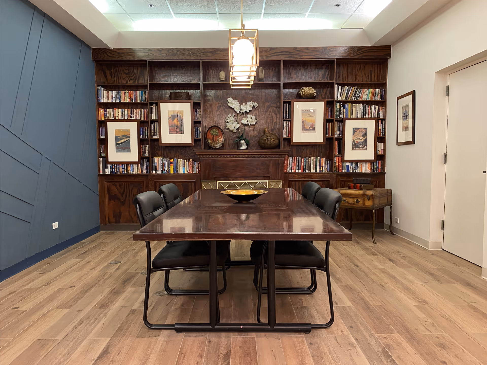 A room with a long wooden table surrounded by six black chairs. Behind the table is a large built-in wooden bookshelf filled with books and framed artwork. The room has light wood flooring, a blue accent wall on the left, and a white wall with a door on the right. A modern pendant light hangs above the table.