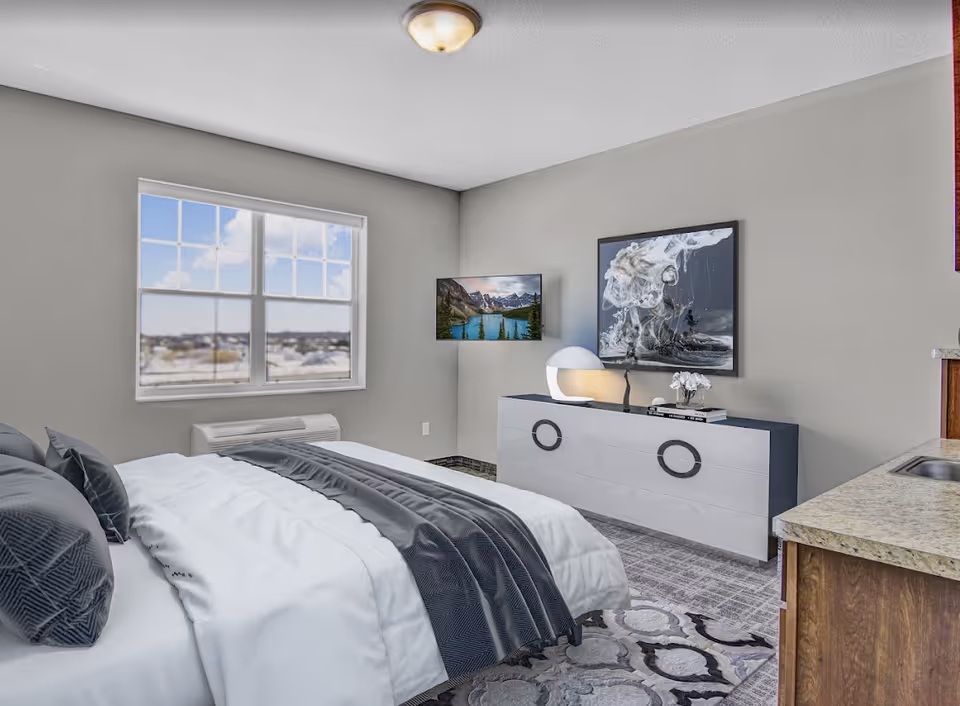 A bright bedroom with a large window showing a cloudy sky outside. The room features a neatly made bed with white and gray bedding, a modern dresser with circular handles, a decorative lamp, a small flower arrangement, and a framed abstract artwork on the wall. A flat-screen TV is mounted on the wall near the corner. Part of a kitchen counter with a sink is visible on the right side.