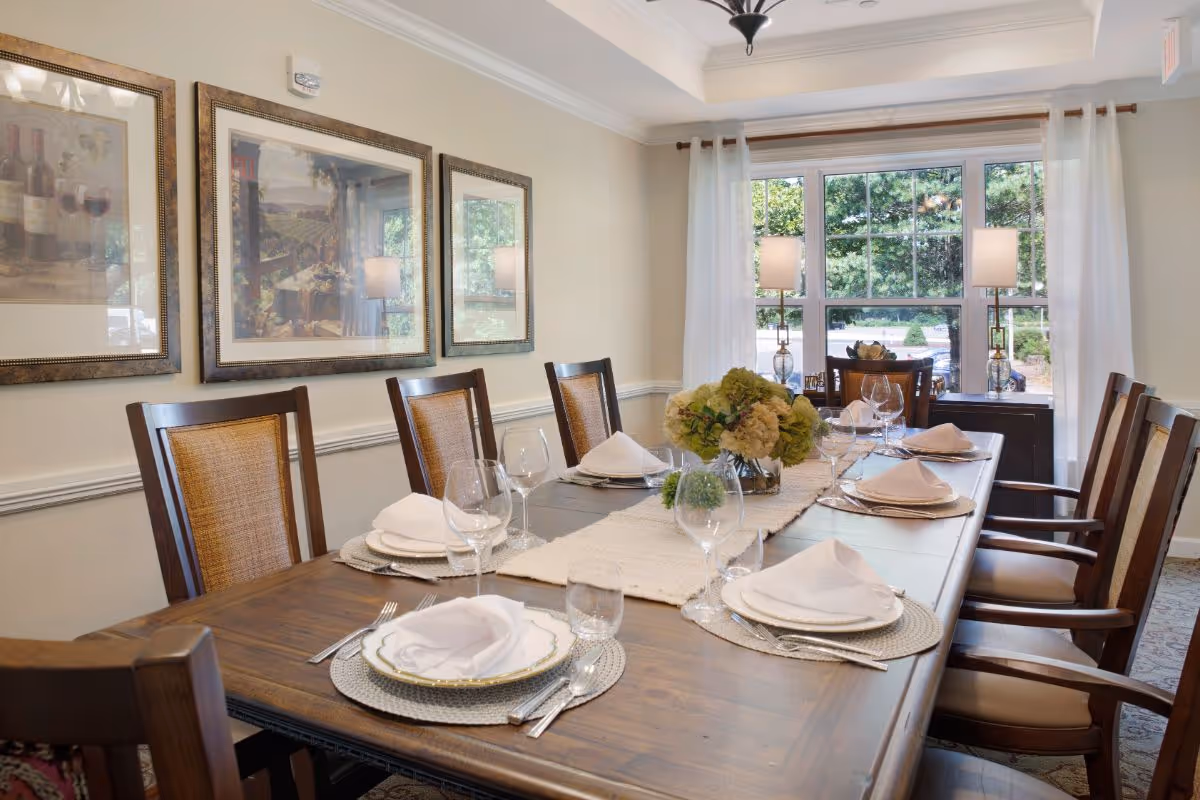 Formal dining room with a long wooden table set with plates, napkins and glassware, surrounded by chairs and a window with lamps.