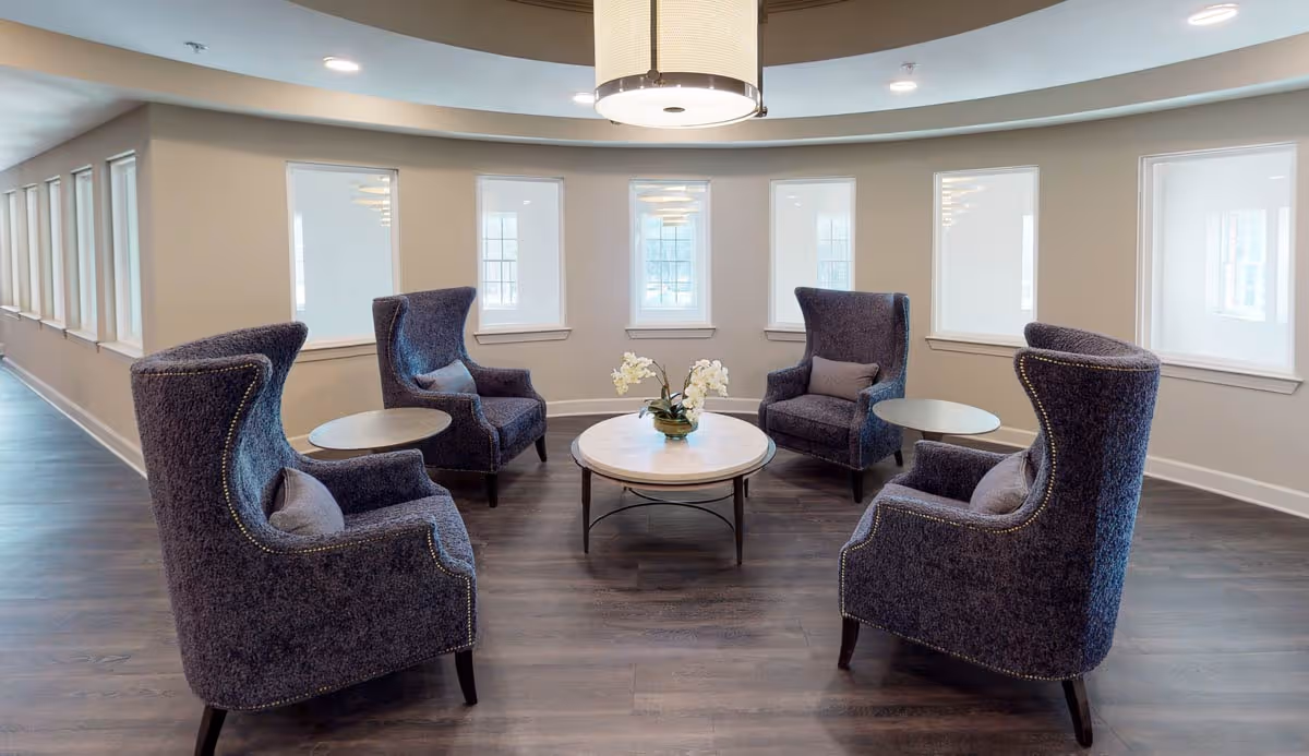 A cluster of high-back upholstered armchairs arranged around a round coffee table beneath a pendant light in a bright curved interior with multiple windows.