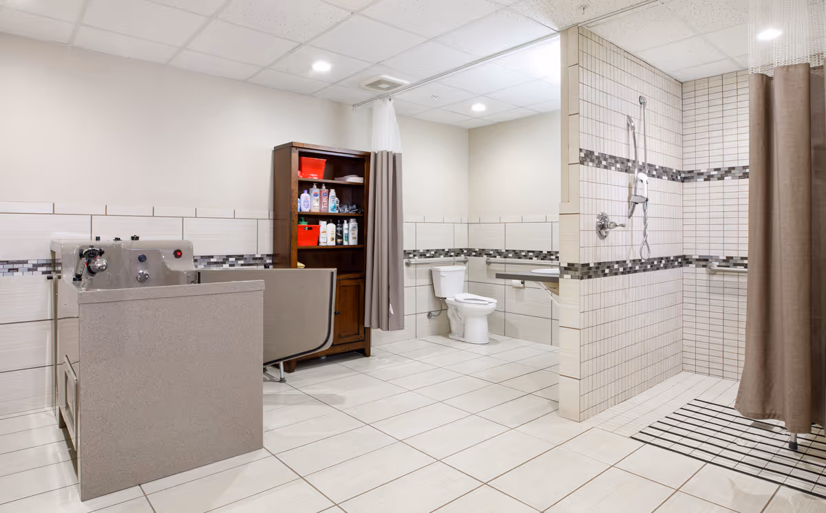 Accessible tiled bathroom with a walk-in shower, toilet, shower lift/chair, and a storage cabinet with supplies.