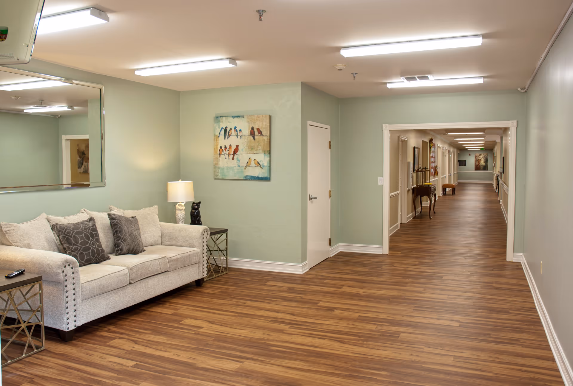 A well-lit interior hallway of a senior living community with light green walls and wood flooring. On the left side, there is a beige sofa with three patterned cushions, a side table with a lamp and a small black cat figurine. A large mirror hangs above the sofa, and a painting of birds is on the wall. The hallway extends into the distance with more doors and framed artwork visible.