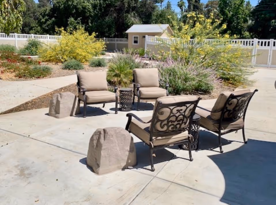 Outdoor patio area with four cushioned metal chairs arranged around a small table on a concrete surface. Surrounding the patio are landscaped garden beds with various shrubs and plants, and a white fence encloses the area. A small shed or structure is visible in the background.