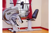 A SciFit exercise machine with a seat and pedals positioned in a room with wooden flooring, cream-colored walls, and windows with red curtains.