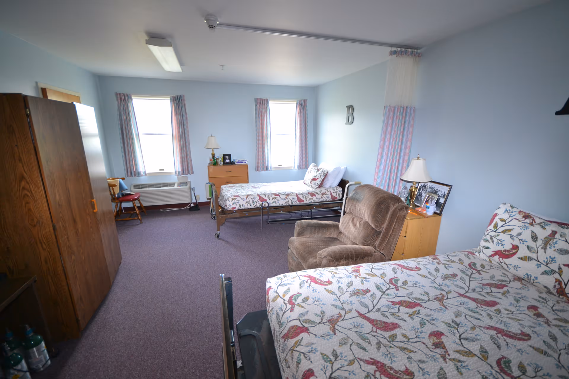 A spacious assisted living bedroom with two beds covered in floral patterned bedspreads, a brown recliner chair, a wooden wardrobe, a small wooden dresser with a lamp and framed photos, two windows with curtains, and light blue walls.