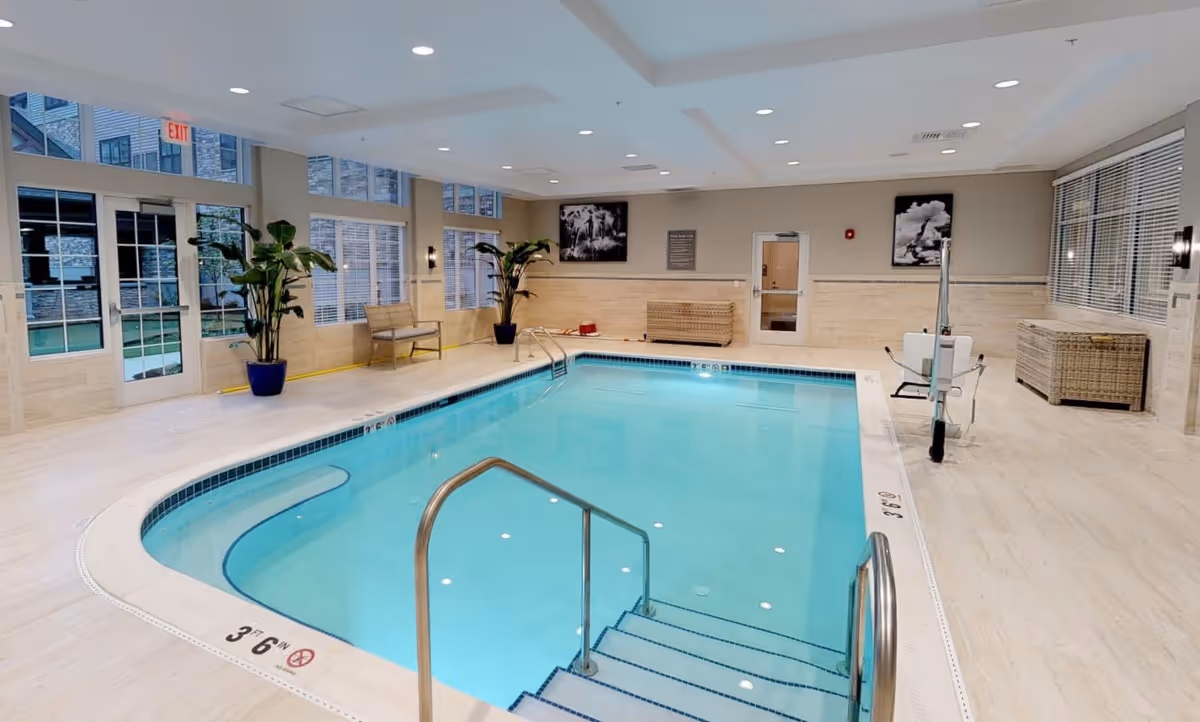 Indoor swimming pool area with clear blue water, metal handrails, steps leading into the pool, potted plants, benches, wicker storage chests, large windows, and bright ceiling lights.