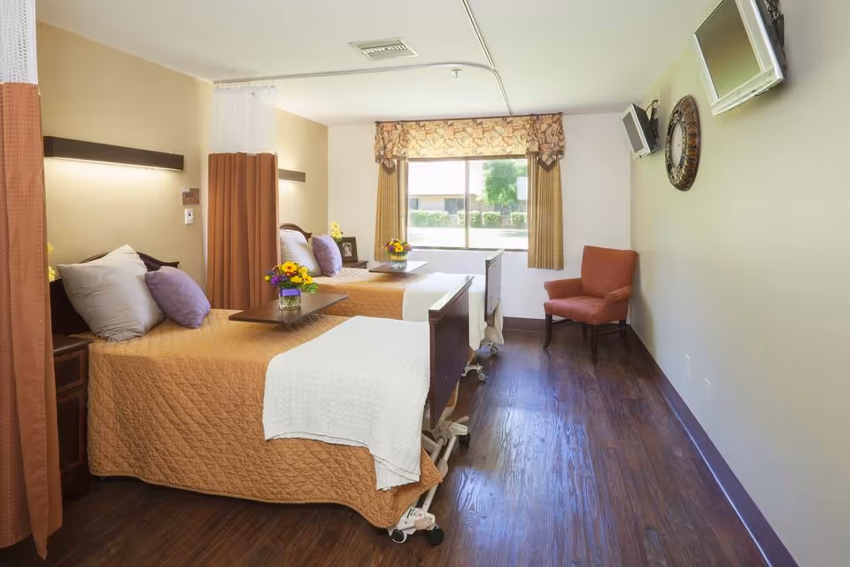 A bright shared patient room with two adjustable beds, overbed trays with flowers, a window, wall-mounted TVs, and a chair.