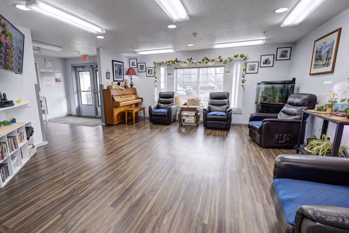 A bright and spacious common area in a senior living facility with wood flooring, several black leather recliners with blue cushions, a wooden piano with a stool, a large window decorated with a garland of green leaves, framed pictures on the walls, a fish tank, and a small table with plants.