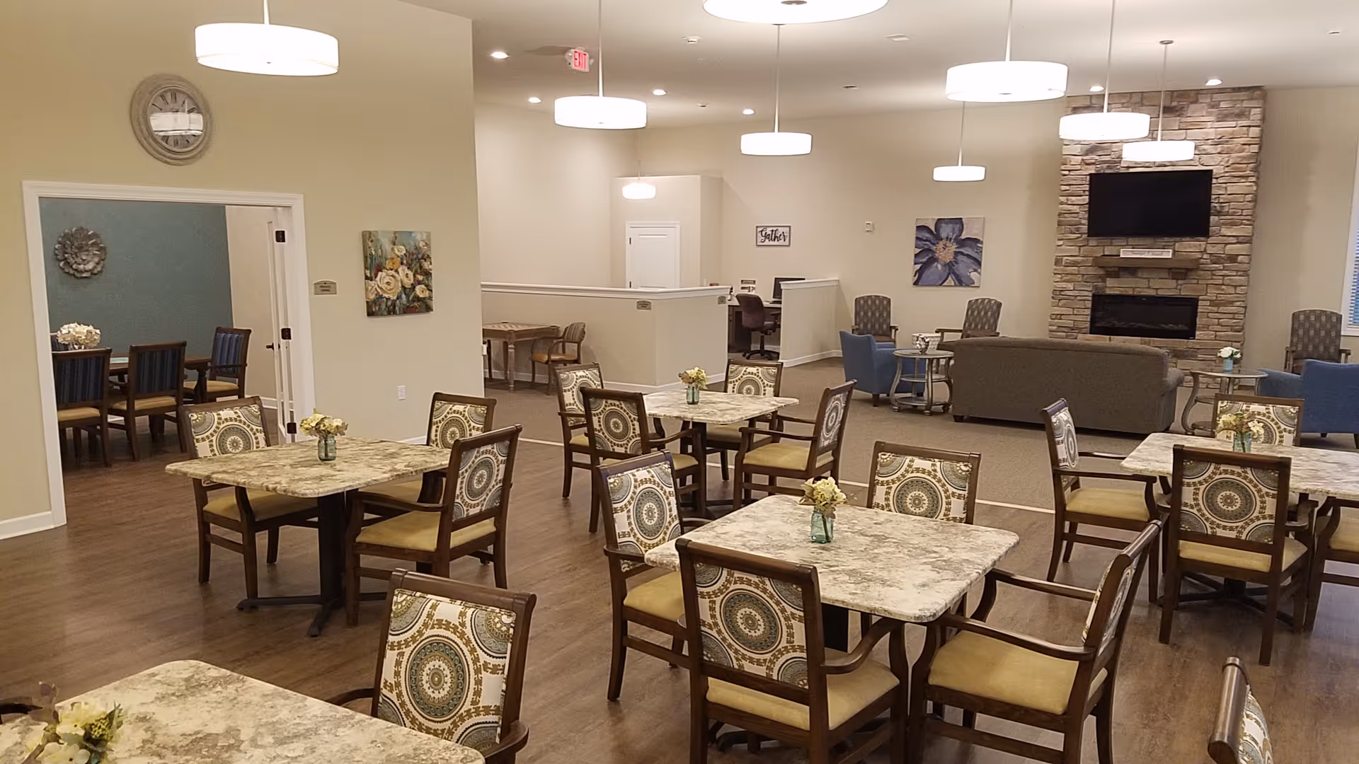 A spacious and well-lit common area in a senior living facility with multiple square tables, each surrounded by four patterned chairs. Small flower arrangements are placed on each table. In the background, there is a seating area with sofas and armchairs arranged around a stone fireplace with a mounted flat-screen TV above it. The walls are decorated with floral artwork, and pendant lights hang from the ceiling. A clock is mounted on the wall near a doorway leading to another room with a dining table and chairs.