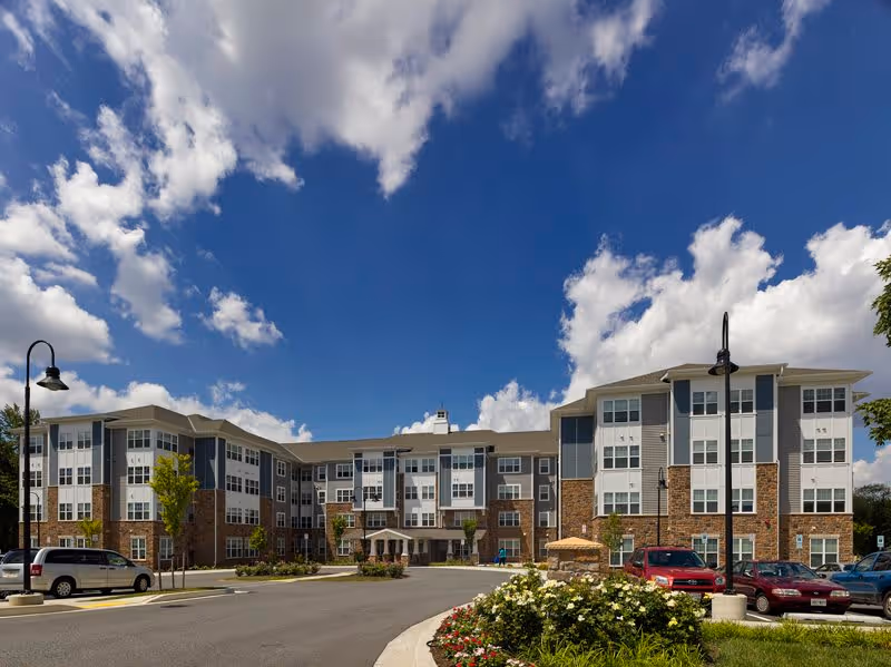 Exterior view of a large, modern senior living facility building under a partly cloudy blue sky. The building has multiple stories with a combination of brick and siding, numerous windows, and a main entrance with a covered drop-off area. There are several cars parked in front and landscaped flower beds near the driveway.